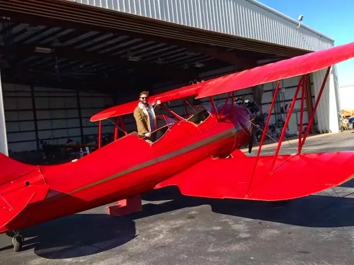 Dan Sloat standing in the cockpit of a red biplane outside a hangar