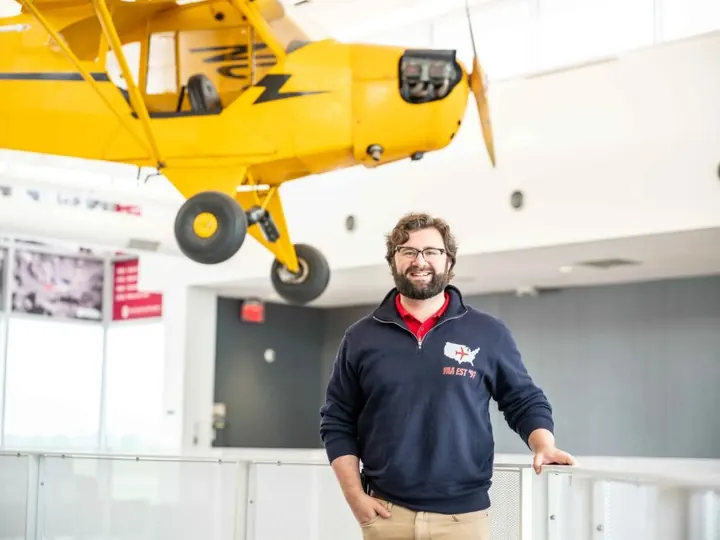 Dan Sloat in a Youth Aviation Adventure sweatshirt below a yellow Piper Cub hanging from a museum ceiling