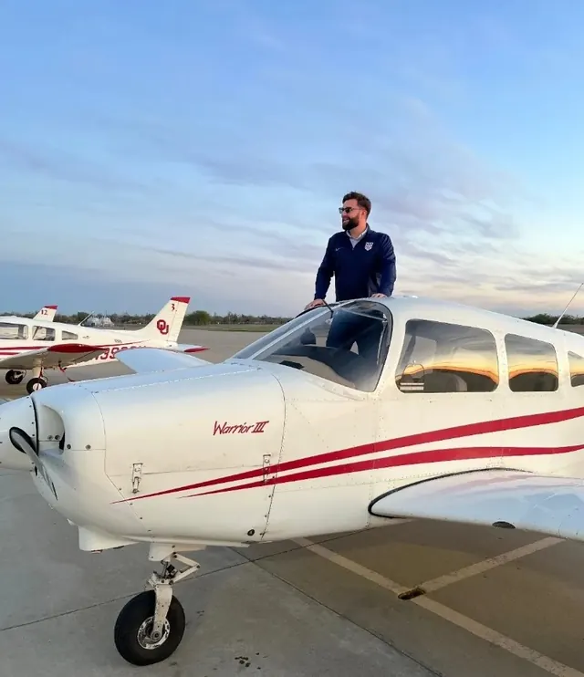 Dan Sloat standing on the wing of a white and red Piper Warrior on the ramp at sunset