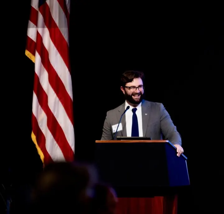 Dan Sloat smiling at a podium on a dark stage beside the American flag