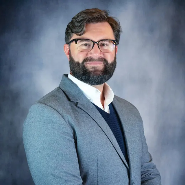 Dan Sloat smiling in a grey blazer, navy sweater and white shirt against a mottled studio backdrop