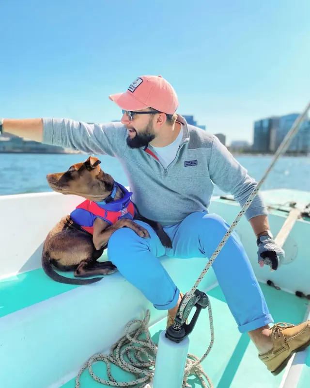 Dan Sloat sailing on Boston Harbor in a pink cap with his dog Deacon in a life jacket